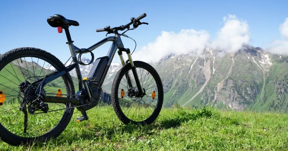 Grey hardtail electric mountain bike with mid-drive motor and frame-mounted battery parked on a grassy alpine ridge, featuring a background of green and rocky mountain peaks under a blue sky.